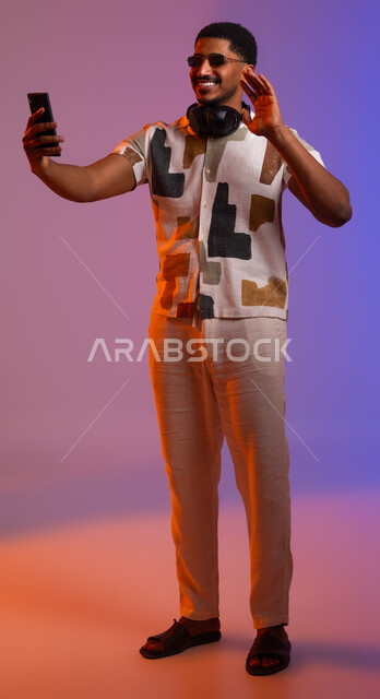 Placing advanced headphones around the neck, broadcasting live to friends via social media, portrait of a smiling Saudi Arabian Gulf man wearing casual summer clothes and sunglasses making a video call via his mobile phone, full length body portrait, colored background