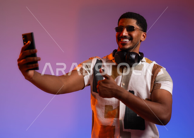 عرض البرامج والتطبيقات المتطورة،, بورتريه مقرب لرجل عربي سعودي خليجي مبتسم يرتدي الزي الكاجوال الصيفي والنظارات الشمسية يضع سماعات الهيدفون حول رقبته يقوم بالتقاط صور سيلفي باستخدام الهاتف المحمول، توثيق اللحظات السعيدة باستخدام الهاتف المحمول, خلفية ملونة