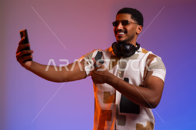 توثيق اللحظات السعيدة باستخدام الهاتف المحمول, بورتريه مقرب لرجل عربي سعودي خليجي مبتسم يرتدي الزي الكاجوال والنظارات الشمسية يضع سماعات الهيدفون على رقبته يقوم بالتقاط صور سيلفي عبر الجوال، عرض البرامج والتطبيقات المتطورة، خلفية ملونة.