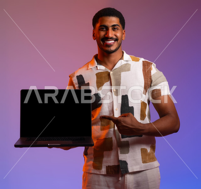 عرض تطبيقات ومواقع الحاسب، متابعة اخبار وسائل التواصل الاجتماعي الكترونيا، بورتريه مقرب لرجل عربي سعودي خليجي مبتسم يرتدي الزي الكاجوال ويشير بإصبعه الى الحاسوب المحمول بشاشة سوداء فارغة، خلفية ملونة