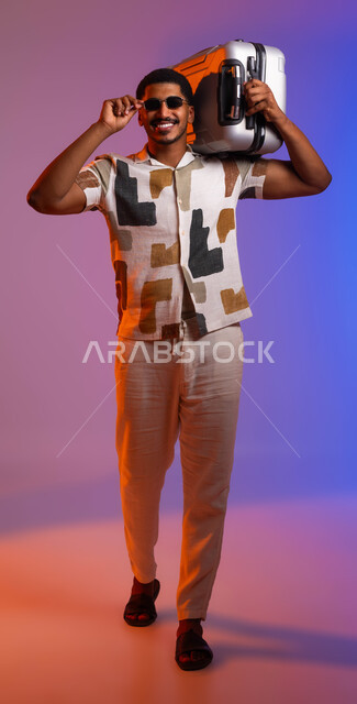 قضاء أوقات ممتعة خارج البلاد، بورتريه لرجل عربي سعودي خليجي مبتسم يرتدي قميص صيفي يضع النظارة الشمسية ويحمل حقيبة السفر، مفهوم السياحة والسفر، ايماءات الحماس لرحلة ممتعه، صورة الجسم بالطول الكامل، خلفية ملونة