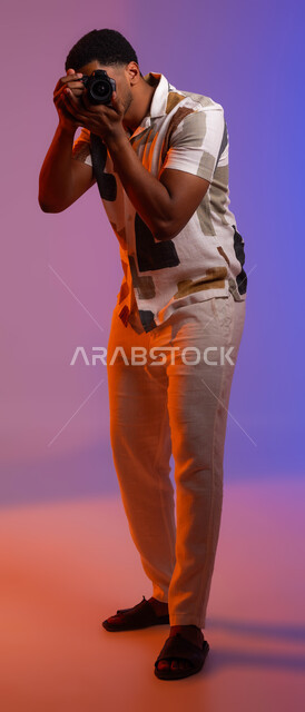 A professional photographer preparing to take pictures, a portrait of a young Saudi Gulf Arab man wearing a casual outfit, holding a camera in his hand, high-quality artistic shots, using modern devices and equipment, full-length body portrait, colored background