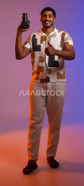 إيماءات القبول والرضا, استخدام الأجهزة والتقنيات الحديثة, بورتريه لرجل عربي خليجي سعودي مبتسم يرتدي الزي الكاجوال يحمل بيده كاميرا عالية الدقة، رفع اصبع الإبهام نحو الأعلى بعلامة الجودة, التقاط الصور التذكارية, صورة الجسم بالطول الكامل, خلفية ملونة .