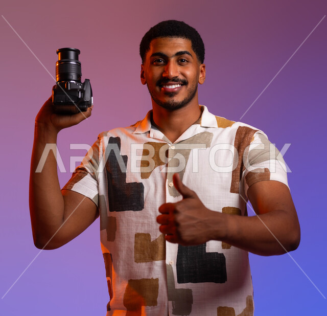 Using modern, high-resolution techniques to take photographs, a portrait of a smiling Saudi Gulf Arab young man wearing a casual outfit, holding a camera in his hand, raising his thumb upward with the quality sign,  documenting happy moments, a colorful background