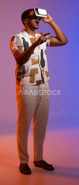 Clicking and pointing at something using the sense of touch, interacting and merging with virtual reality games, a close-up portrait of a Saudi Gulf Arab man wearing casual clothes and 3D smart glasses, gestures of enjoyment and focus in playing, spending a fun summer vacation, colorful background