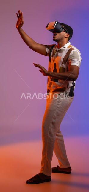 Simulating the imaginary world and trying to touch an item, gestures for interaction and integration with virtual reality games, a close-up portrait of a Saudi Gulf Arab man wearing casual clothes and 3D smart glasses, using modern devices and technologies, full-length body image, colored background