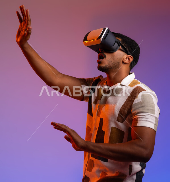 إستخدام حاسة اللمس لتحريك العناصر الهولوجرامية ، بورتريه لرجل عربي خليجي سعودي يرتدي الزي الكاجوال ونظارات الواقع الإفتراضي (في آر) ثلاثية الأبعاد ,  مفهوم التقنيات الإلكترونية الحديثة، محاولة لمس شيء ما , خلفية ملونة .