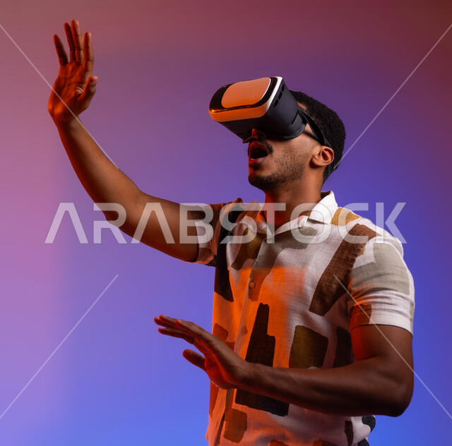 التفاعل والاندماج مع الواقع الافتراضي , محاكاة العالم الخيالي ومحاولة لمس عنصر ما , بورتريه مقرب لشاب عربي سعودي خليجي يرتدي الزي الكاجوال و النظارات الذكية ثلاثية الأبعاد, إيماءات الاستمتاع والتركيز, قضاء عطلة صيفية ممتعة , خلفية ملونة .