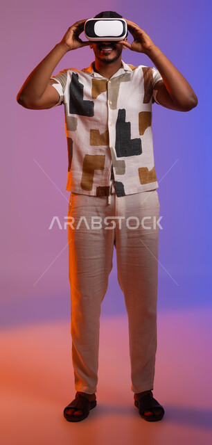 التسلية والترفيه مع التجارب الحية وألعاب الواقع الافتراضي في العطلة الصيفية , مفهوم التكنولوجيا والأجهزة الحديثة , بورتريه لشاب عربي سعودي خليجي مبتسم يرتدي الزي الكاجوال و النظارات الذكية ثلاثية الأبعاد, صورة الجسم بالطول الكامل, خلفية ملونة .