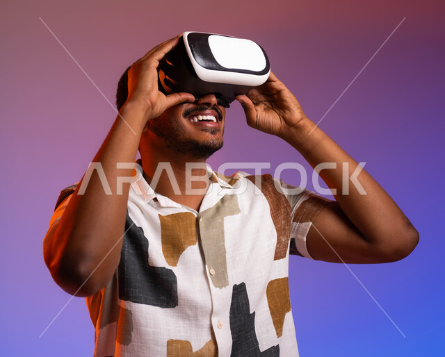Concept of modern devices and technologies, close-up portrait of a smiling Saudi Arabian Gulf man wearing casual clothes and 3D smart glasses, exploring tourist destinations through virtual reality glasses, gestures of happiness and enjoyment on summer vacation, colorful background
