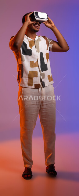 الاندماج مع الأفلام ومحاكاة العالم الخيالي، بورتريه لرجل عربي سعودي خليجي مبتسم يرتدي الزي الكاجوال والنظارات الذكية ثلاثية الأبعاد, قضاء أوقات ممتعة في الإجازة مع ألعاب الواقع الافتراضي, صورة الجسم بالطول الكامل , خلفية ملونة .