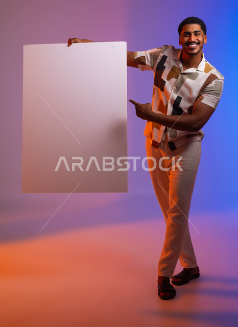 التسويق والترويج لشيء ما، موك اب أبيض، النظر إلى الكاميرا بإيماءات الإعجاب والسرور, بورتريه لشاب عربي سعودي خليجي مبتسم يرتدي الزي الكاجوال يحمل بيده لوحه بيضاء مستطيلة، الإشارة إلى لوح إعلاني فارغ، إستخدام لوحات توضيحية ، خلفية ملونة