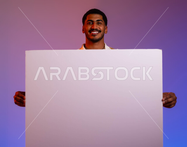 النظر الى الكاميرا بإيماءات الإعجاب والسرور، موك اب أبيض، بورتريه مقرب لشاب عربي سعودي خليجي مبتسم يحمل بيده لوحه بيضاء مستطيلة، لوح إعلاني فارغ، الدعاية والترويج لشيء ما ، خلفية ملونة