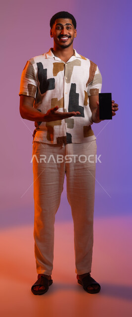 استخدام الأجهزة والتقنيات الحديثة في السفر, بورتريه لرجل عربي خليجي سعودي مبتسم يرتدي الزي الكاجوال يعرض تطبيقات الهاتف المحمول , بث مباشر وإجراء مكالمات فيديو, التقاط الصور التذكارية في الرحلات الصيفية السعيدة , صورة الجسم بالطول الكامل, خلفية ملونة