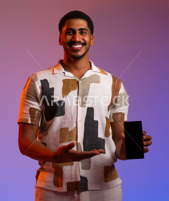 تطبيقات الدليل السياحي عبر الأجهزة التقنية الحديثة , توثيق لحظات العطلة السعيدة باستخدام الهاتف المحمول, البحث عن المواقع السياحية الترفيهية عبر الانترنت ,بورتريه لرجل عربي خليجي سعودي مبتسم يرتدي الزي الكاجوال يشير إلى شاشة سوداء فارغة, خلفية ملونة .