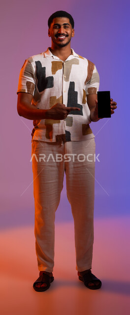 الإشارة إلى شاشة سوداء فارغة, متابعة العروض والتخفيضات للرحلات البحرية عبر تطبيقات التواصل الاجتماعي , بورتريه لرجل عربي خليجي سعودي مبتسم يرتدي الزي الكاجوال ويحمل الهاتف المحمول , خدمات التجوال والأنشطة السياحية, صورة الجسم بالطول الكامل, خلفية ملونة .