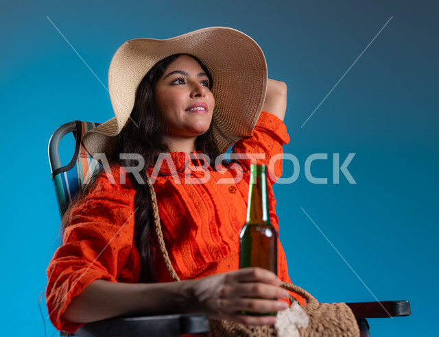 الاسترخاء بوقت الفراغ في عطلة الصيف، بورتريه لفتاة عربيه خليجية سعودية مبتسمة ترتدي الزي الكاجوال وقبعة القش الشمسية تجلس على الكرسي وتنظر الى الاعلى، الاستمتاع بالمشروبات الغازيه المنعشة، ايماءات التأمل والراحة، خلفية زرقاء