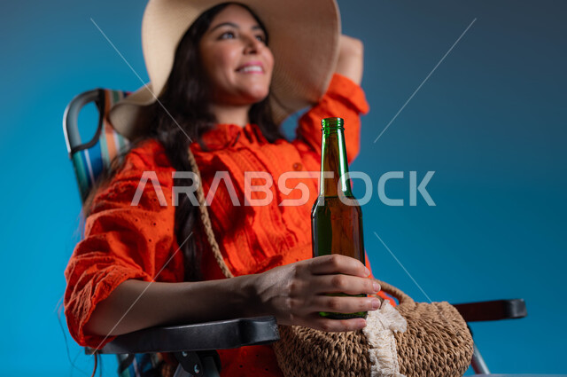 الاستمتاع بالمشروبات الصيفة المنعشة، بورتريه مقرب لإمرأة عربيه خليجية سعودية مبتسمه ترتدي الزي الكاجوال وقبعة القش العصرية تنظر إلى الأعلى، الجلوس على الكرسي والاستمتاع بعطلة الصيف، ايماءات التأمل والسرور، خلفية زرقاء