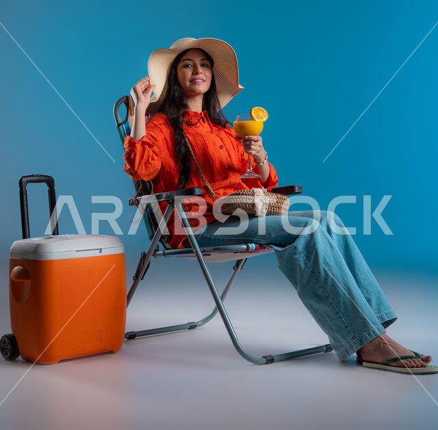 الانتعاش بالمشروبات الباردة في فصل الصيف، بورتريه لسيدة عربيه خليجية سعودية ترتدي الزي الكاجوال وقبعة القش العصرية تقوم بتعديل القبعة بجانبها براد الرحلات، الجلوس على الكرسي للراحة والاسترخاء، متعة السفر و الرحلات السياحية، صورة لكامل الجسم، خلفية زرقاء