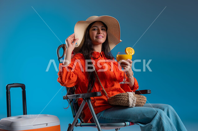 Spending fun times on tourist trips during the summer vacation, a portrait of a young Saudi Arabian Gulf woman wearing a casual outfit and a straw hat, holding the hat in her hand with a travel cooler next to her, the concept of preparing and preparing for travel, enjoying a cold orange juice drink, blue background.