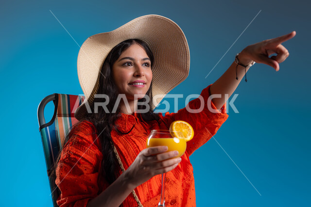 الإستعداد لرحلة سياحية في الإجازة الصيفية، بورتريه لسيدة عربيه خليجية سعودية مبتسمه ترتدي الزي الكاجوال وقبعة القش العصرية تشير إلى شيء ما باصبع السبابة، الاستمتاع بمشروب عصير البرتقال الطازج، ايماءات السرور والراحة في العطلة، خلفية زرقاء.