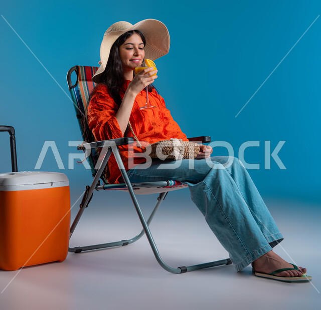 الهروب من حرارة الصيف باحتساء المشروبات الباردة، الجلوس على الكرسي للاسترخاء، بورتريه لشابة عربيه خليجية سعودية مبتسمه ترتدي الزي الكاجوال و قبعة القش العصرية بجانبها كولر الرحلات، ايماءات الراحة والاستمتاع، صورة الجسم بالطول الكامل، خلفية زرقاء.