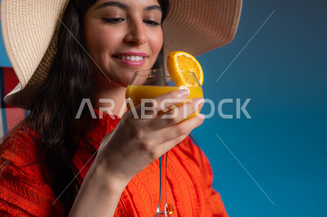 قضاء أوقات ممتعة و الابتعاد عن الروتين اليومي، بورتريه مقرب لشابة عربية خليجية سعودية ترتدي الزي الكاجوال وتضع القبعة العصرية تستمتع بشرب كأس البرتقال البارد في الأجواء الصيفية، رحلة سياحية في الإجازة، إيماءات الاسترخاء والترفيه عن النفس، خلفية زرقاء.