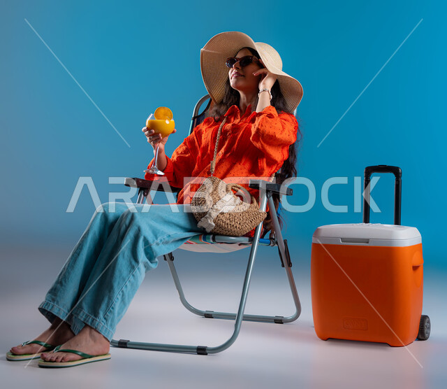 مفهوم الجلوس والراحة وقضاء عطلة صيفية سعيدة، بورتريه لإمرأة عربيه خليجية سعودية مبتسمه ترتدي الزي الكاجوال وتضع قبعة القش والنظارة الشمسية وبجانبها براد الرحلات تنظر للأعلى، الاستمتاع بالمشروبات البارده في الاجواء الحاره، صورة الجسم بالطول الكامل، خلفية زرقاء.