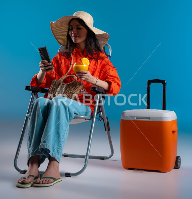 تصفح مواقع التواصل الاجتماعي بإيماءات السرور، بورتريه لامرأة عربية سعودية خليجية ترتدي اللباس الكاجوال وقبعة القش بجانبها حافظة تبريد تقوم بمتابعة آخر الأخبار، الاستمتاع بعصير البرتقال الطازج، استخدام التقنيات المتطورة، صورة الجسم بالطول الكامل، خلفية زرقاء.