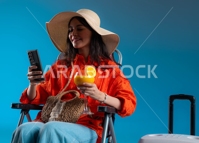 التواصل عن بعد عبر وسائل التواصل الاجتماعي، بورتريه لشابة عربية خليجية سعودية مبتسمة ترتدي الزي الكاجوال وقبعة القش تجلس على الكرسي وتقوم بتصفح الإنترنت عبر الجوال، قضاء أوقات ممتعة في ألعاب الاونلاين ، مفهوم التسوق الالكتروني ، خلفية زرقاء.