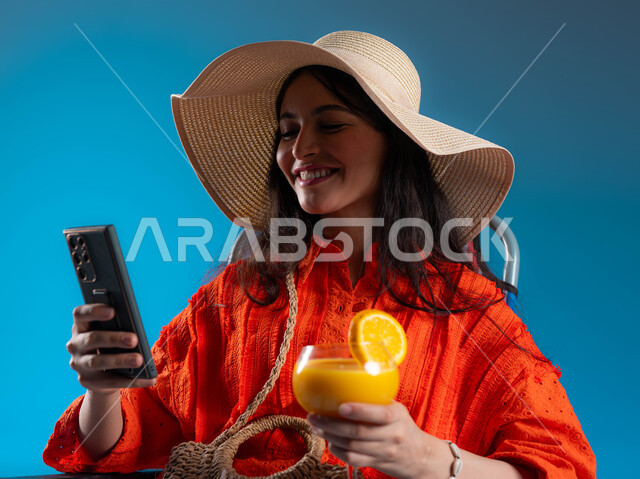 استخدام جهاز الهاتف المحمول و إيماءات تدل على السعادة، بورتريه لسيدة عربية سعودية خليجية ترتدي الزي الكاجوال و قبعة القش الشمسية المعاصرة، الاستمتاع بشراب البرتقال الطبيعي و الدردشة على الهاتف، مفهوم تطوير العلاقات الإجتماعية، خلفية زرقاء.