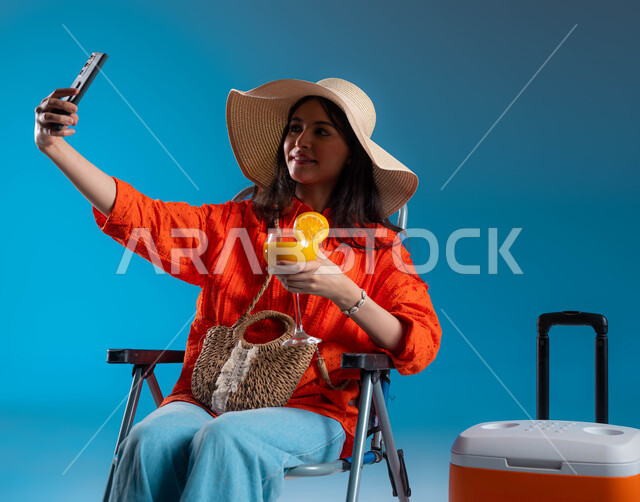 المتعة بالتقاط صورة سيلفي من خلال الهاتف المحمول، بورتريه لشابة عربية سعودية خليجية ترتدي الزي الكاجوال و قبعة القش العصرية بجانبها برادة الرحلات، الراحة والجلوس على كرسي، توثيق الذكريات في الرحلات السياحية، الاسمتاع بمشروب البرتقال البارد، خلفية زرقاء.