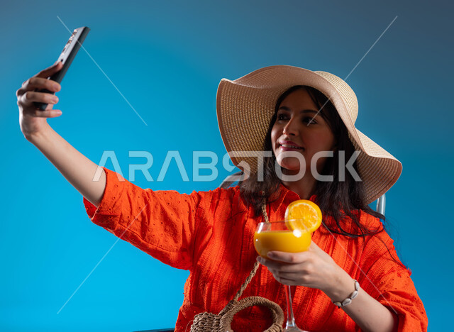 التقاط صور سيلفي تذكارية باستخدام الهاتف المحمول ، بورتريه لسيدة عربية سعودية خليجية ترتدي الزي الكاجوال والقبعة القش الصيفية ، الاستمتاع بعصير البرتقال البارد في الأجواء الحارة ، لجلوس على الكرسي والترفيه في الخارج ، هواية وفنون التصوير ، خلفية زرقاء.