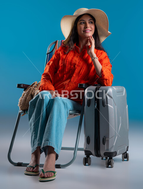 عطلة صيفية مليئة بالرحلات الممتعة، بورتريه لامرأة عربية سعودية خليجية ترتدي الملابس الكاجوال و قبعة القش المعاصرة بجانبها حقيبة سفر، استغلال الإجازات في السفر، صورة الجسم بالطول الكامل، الجلوس على كرسي، ، خلفية زرقاء .