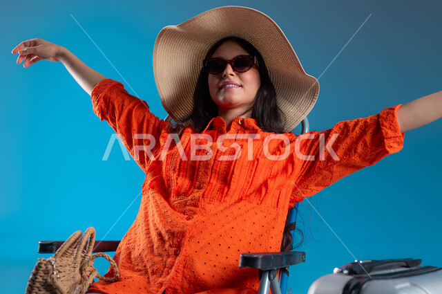 الاستعداد لقضاء العطلة الصيفية ، بورتريه لشابة سعودية عربية خليجية مبتسمة ترتدي لباس كاجوال تضع قبعة القش والنظارة الشمسية و تجلس على كرسي وتفتح ذراعيها بسرور و انشراح ، إيماءات الاسترخاء و الهواء الطلق ، مفهوم السياحة والسفر ،خلفية زرقاء.