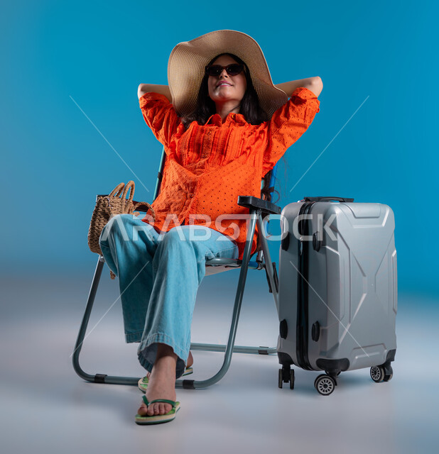 الجلوس والاستمتاع بالإجازة والمناظر الطبيعيه, الرحلات السياحية في إحدى المنتجعات والشواطئ السعودية, بورتريه لشابة عربية خليجية سعودية مبتسمة ترتدي الزي الكاجوال والنظارة الشمسية مع قبعة القش ترفع يديها للأعلى بإيماءات الاسترخاء والراحة, خلفية زرقاء