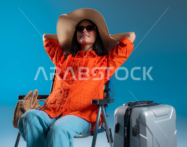 الجلوس والاستمتاع بالإجازة والمناظر الطبيعيه, الرحلات السياحية في إحدى المنتجعات والشواطئ السعودية, بورتريه لشابة عربية خليجية سعودية مبتسمة ترتدي الزي الكاجوال والنظارة الشمسية مع قبعة القش ترفع يديها للأعلى بإيماءات الاسترخاء والراحة, خلفية زرقاء