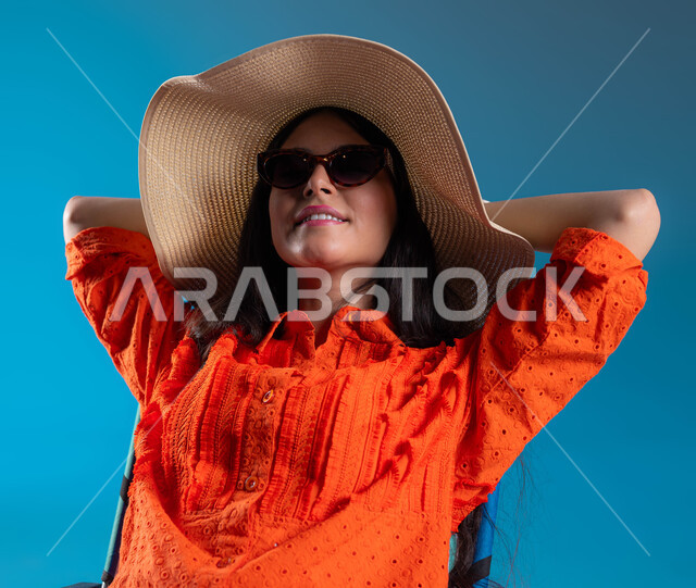 الجلوس والاستمتاع بالإجازة والمناظر الطبيعيه, الرحلات السياحية في إحدى المنتجعات والشواطئ السعودية, بورتريه لشابة عربية خليجية سعودية مبتسمة ترتدي الزي الكاجوال والنظارة الشمسية مع قبعة القش ترفع يديها للأعلى بإيماءات الاسترخاء والراحة, خلفية زرقاء