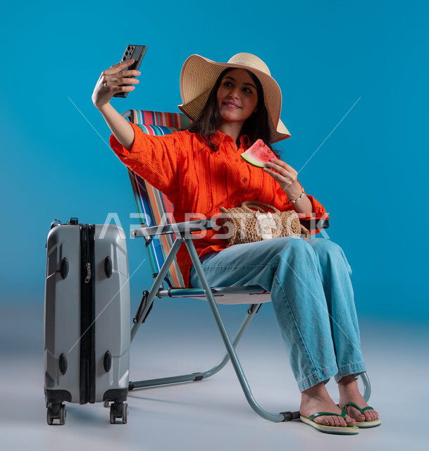 قضاء أيام العطلة بالأماكن الترفيهيه, التقاط صور سيلفي تذكارية عبر الجوال, الاستمتاع بتناول الفواكه الصيفية المنعشة, بورتريه لشابة عربية خليجية سعودية مبتسمة ترتدي الزي الكاجوال وقبعة القش تقوم بإجراء مكالمة فيديو عبر الهاتف المحمول, خلفية زرقاء