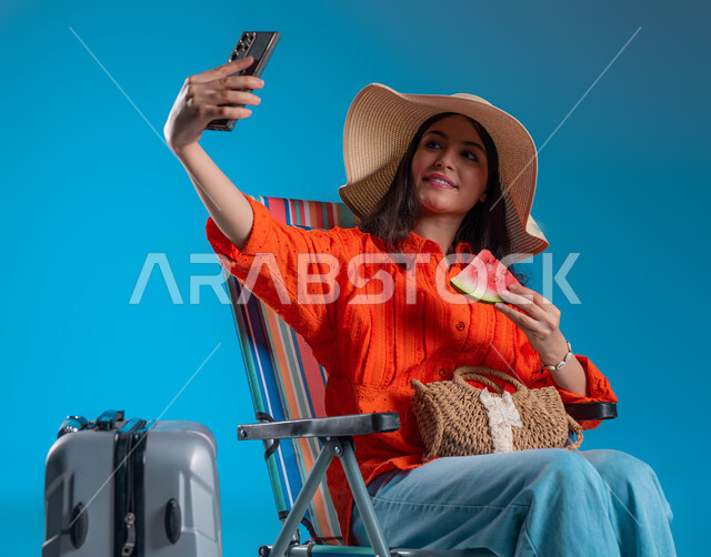 Spending vacation days in entertainment places, taking souvenir selfies via mobile phone, enjoying refreshing summer fruits, close-up portrait of a smiling Saudi Gulf Arab woman wearing a casual outfit and a straw hat making a video call via mobile phone, blue background