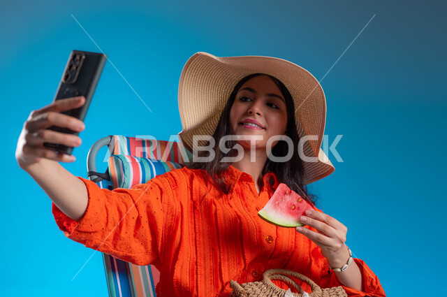 قضاء أيام العطلة بالأماكن الترفيهيه, التقاط صور سيلفي تذكارية عبر الجوال, الاستمتاع بتناول الفواكه الصيفية المنعشة, بورتريه مقرب لشابة عربية خليجية سعودية مبتسمة ترتدي الزي الكاجوال وقبعة القش تقوم بإجراء مكالمة فيديو عبر الهاتف المحمول, خلفية زرقاء