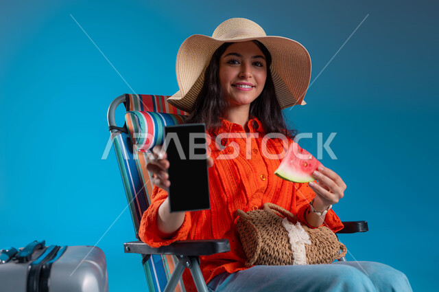 عرض تطبيقات وخدمات السياحة والسفر، الاستعداد للسفر الممتع, الاستمتاع بتناول الفواكه الصيفية المنعشة, بورتريه مقرب لشابة عربية خليجية سعودية مبتسمة ترتدي الزي الكاجوال وقبعة القش تمسك الهاتف المحمول بشاشة سوداء فارغة, خلفية زرقاء.