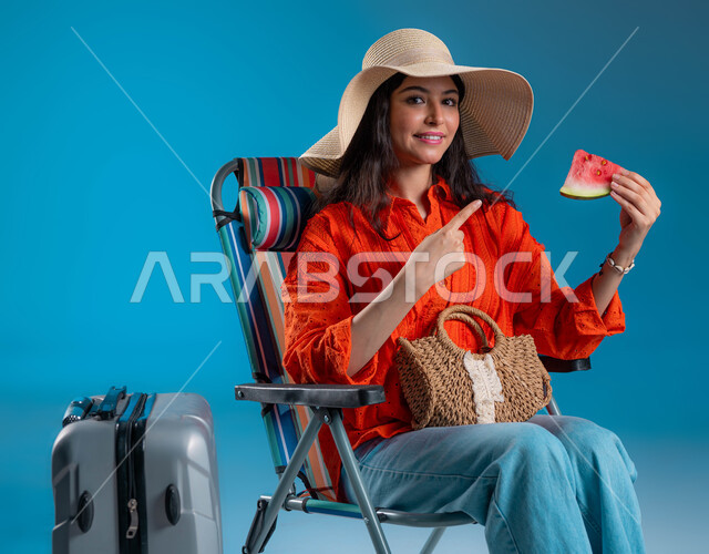 التسلية والترفيه في الخارج، الاستعداد للسفر الممتع, الاستمتاع بتناول الفواكه الصيفية المنعشة, بورتريه لشابة عربية خليجية سعودية مبتسمة ترتدي الزي الكاجوال وقبعة القش تشير بيدها إلى قطعة الفاكهة بإيماءات السرور والمتعة, خلفية زرقاء.