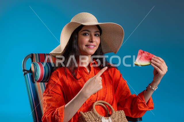 التسلية والترفيه في الخارج، الاستعداد للسفر الممتع, الاستمتاع بتناول الفواكه الصيفية المنعشة, بورتريه مقرب لشابة عربية خليجية سعودية مبتسمة ترتدي الزي الكاجوال وقبعة القش تشير بيدها إلى قطعة الفاكهة بإيماءات السرور والمتعة, خلفية زرقاء.