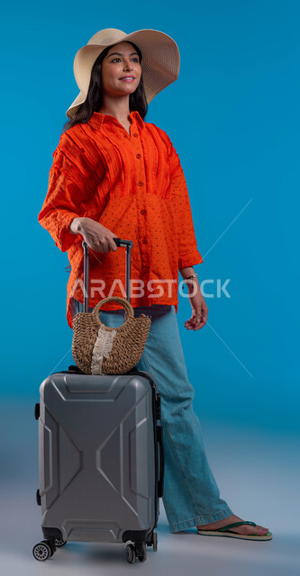 الاستمتاع بإجازة الصيف والهروب من ضغوط الحياة، الاستعداد للسفر الممتع, الاهتمام بالموضة والأزياء العصرية، بورتريه لشابة عربية خليجية سعودية مبتسمة ترتدي الزي الكاجوال و قبعة القش تمسك حقيبة الأمتعة بإيماءات السرور والمتعة, خلفية زرقاء.