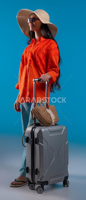 الاستمتاع بإجازة الصيف والهروب من ضغوط الحياة، الاستعداد للسفر الممتع, الاهتمام بالموضة والأزياء العصرية، بورتريه لشابة عربية خليجية سعودية مبتسمة ترتدي الزي الكاجوال والنظارة الشمسية مع قبعة القش تمسك حقيبة الأمتعة بإيماءات السرور والمتعة, صورة الجسم بالكامل، خلفية زرقاء.