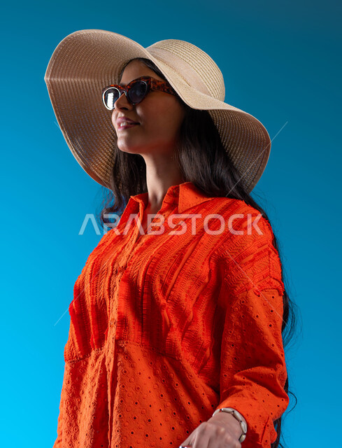 الوقوف بإيماءات الثقة والسعادة والسرور, الاستعداد لقضاء نزهة صيفية ممتعة، مفهوم الأنوثة والأناقة، الاهتمام بالأزياء والموضة العصرية, بورتريه مقرب لشابة عربية خليجية سعودية مبتسمة ترتدي الزي الكاجوال والنظارات الشمسية مع قبعة القش، خلفية زرقاء.