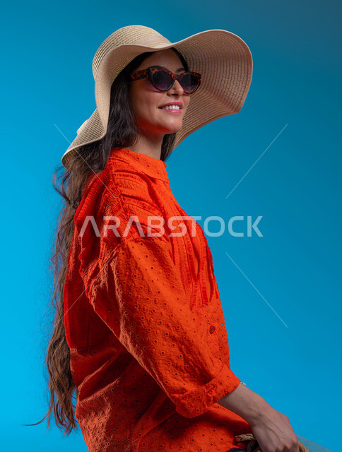 Preparing for a fun summer outing, the concept of femininity and elegance, interest in fashion and modern fashion, a close-up portrait of a smiling Saudi Arabian Gulf woman wearing a casual outfit and sunglasses with a straw hat and looking at something, standing with gestures of confidence, happiness and pleasure, blue background.