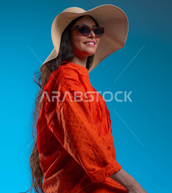 مفهوم الأنوثة والأناقة، الاهتمام بالأزياء والموضة العصرية, بورتريه مقرب لشابة عربية خليجية سعودية مبتسمة ترتدي الزي الكاجوال والنظارات الشمسية مع قبعة القش وتنظر إلى شيء ما، الوقوف بإيماءات الثقة والسعادة والسرور, خلفية زرقاء.