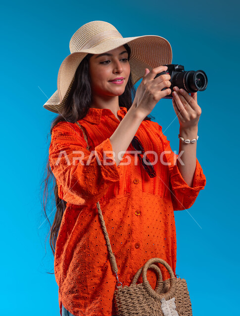 الاستمتاع بتصوير الرحلات الصيفية والأماكن السياحية بالمملكة، استخدام كاميرا التصوير الفوتوغرافي لالتقاط الصور التذكارية وتوثيق اللحظات السعيدة، بورتريه مقرب لشابة عربية خليجية سعودية ترتدي الزي الكاجوال، إيماءات تدل على التركيز، خلفية زرقاء.